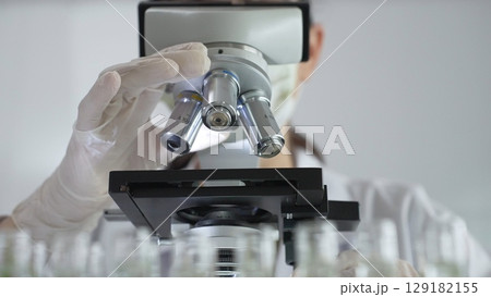 Female scientist wearing white protective gear analyzing test samples under microscope in contemporary research environment, close up of white gloved hand. Medicine and science concept 129182155