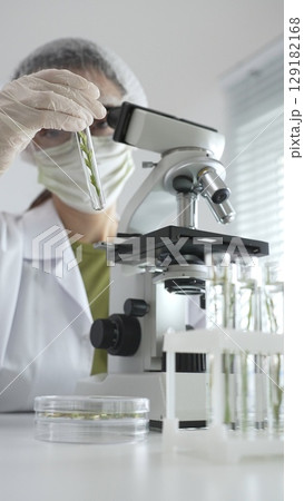 Scientist in white protective mask and gloves holding test tube with plant sample, researching botany and medical applications using a microscope. Medicine, health care and science concept 129182168