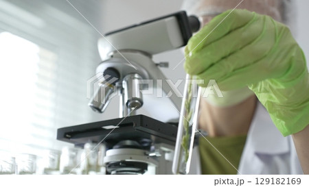 Scientist wearing green gloves and a face mask selecting a test tube with transparent liquid from a rack near a microscope in a lab. Medicine, healthcare and science concept Scientist wearing green gloves and a face mask selecting a test tube with transparent liquid from a rack near a microscope in a lab. Medicine, healthcare and science concept 129182169