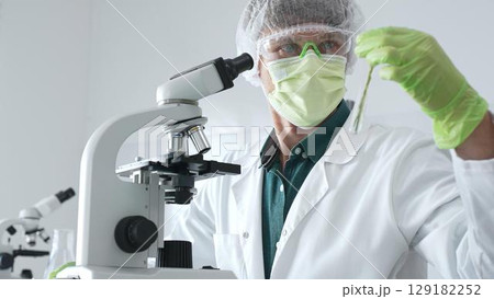 Adult male scientist wearing green protective gear looking at plant samples in test tubes and using a microscope in modern laboratory for botanical research. Medicine, healthcare and science concept Adult male scientist wearing green protective gear looking at plant samples in test tubes and using a microscope in modern laboratory for botanical research. Medicine, healthcare and science concept 129182252