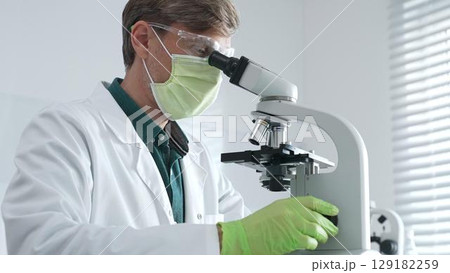 Adult male scientific professional analyzing sample through microscope, wearing green protective gear in sterile laboratory setting. Medicine, healthcare and science concept 129182259