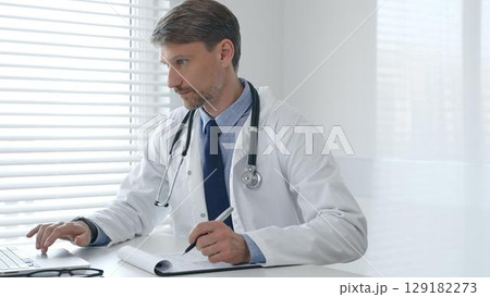 A male doctor in a lab coat with a stethoscope sits at a table in a bright medical office and uses a laptop while taking notes on a clipboard for patient care and treatment 129182273