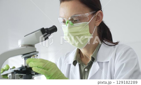 Female scientist examining specimen, placing slide under microscope while wearing green protective gear in sterile laboratory workspace. Medicine, healthcare and science concept 129182289