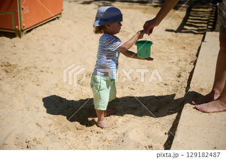 Young Child Playing at the Beach While Holding a Sand Bucket Handed by Adult 129182487