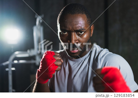 Boxer in gym. Aggressive African man fighter resting after training boxing ready for fight looking at camera. Strong sweated man training punches looking concentrated straight preparing for sparring 129183854