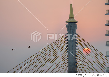 Closeup silhouette of a Bhumibol Bridge with the sun going down over the bridge during orange sunset sky. Closeup silhouette of a Bhumibol Bridge with the sun going down over the bridge during orange sunset sky. 129184249