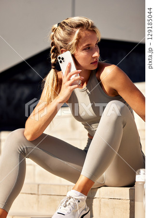 Woman in activewear holding a smartphone while sitting outdoors on sunny steps 129185044