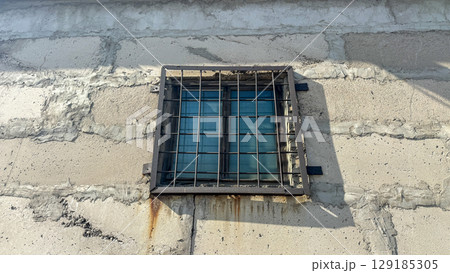 Grungy concrete wall with a small barred window featuring a heavy metal security grate. 129185305
