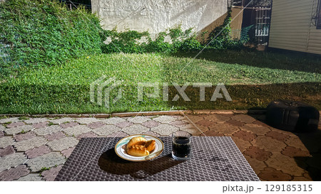 Night snack on outdoor table in backyard garden. A plate with baked pastries and a glass of dark soda on a wicker-style table in a backyard at night. The yard is lit with artificial light. Night snack on outdoor table in backyard garden. A plate with baked pastries and a glass of dark soda on a wicker-style table in a backyard at night. The yard is lit with artificial light. 129185315