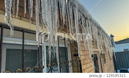 Long glistening icicles hanging from a building roof at sunset, reflecting warm golden light on a cold winter day. Long glistening icicles hanging from a building roof at sunset, reflecting warm golden light on a cold winter day. 129185321