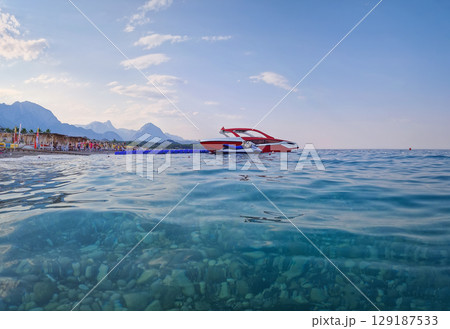 Panoramic shot of Kemer beach with clear turquoise water revealing pebbles below. Speedboats anchored near the shore, and wonderful Taurus mountains in the background under a blue sky Panoramic shot of Kemer beach with clear turquoise water revealing pebbles below. Speedboats anchored near the shore, and wonderful Taurus mountains in the background under a blue sky 129187533