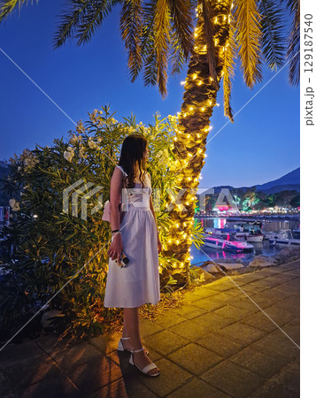 Charming evening scene with a gorgeous woman in a white dress, stands by a palm tree adorned with festive string lights, looking at the vibrant Moonlight beach at night in Kemer, Turkey 129187540