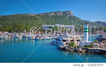 Kemer marina filled with numerous boats and yachts with Calis Hill in the background. A lighthouse stands prominently on the rocky pier, overlooking the clear blue water under a bright blue sky 129187555