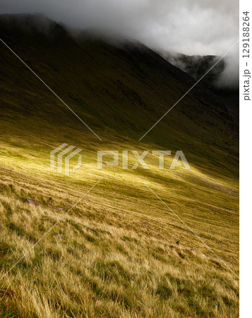 Grassy hillside in Coomloughra Horseshoe, Ireland is bathed in sunlight with areas of shadow, creating textures and contrasting light. 129188264