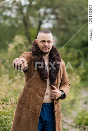 Rugged Man in Brown Suede Coat Pointing Forward in Forest, Nature Outdoor Scene, Dramatic Look, Bearded Male, Winter Fashion, Open Coat. Rugged Man in Brown Suede Coat Pointing Forward in Forest, Nature Outdoor Scene, Dramatic Look, Bearded Male, Winter Fashion, Open Coat. 129188408