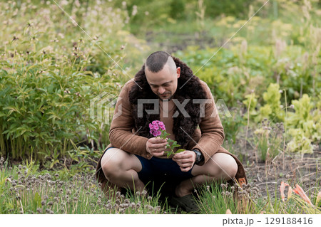 Man Kneeling in Meadow with Pink Flower, Nature Scene, Rustic Brown Fur Coat, Green Field, Soft Lighting, Emotional Moment, Springtime Pastoral. Man Kneeling in Meadow with Pink Flower, Nature Scene, Rustic Brown Fur Coat, Green Field, Soft Lighting, Emotional Moment, Springtime Pastoral. 129188416