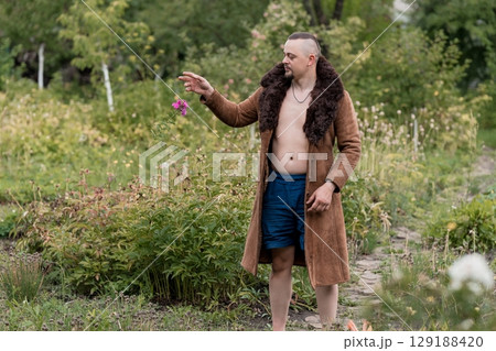 Muscular Man in Robe Holding Flower in Meadow. Nature, Bare Chest, Warm Tone, Outdoor Portrait, Rustic Style, Green Foliage, Summer Day. Muscular Man in Robe Holding Flower in Meadow. Nature, Bare Chest, Warm Tone, Outdoor Portrait, Rustic Style, Green Foliage, Summer Day. 129188420