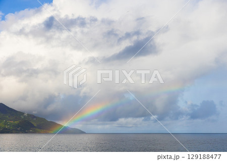 Vibrant Rainbow Over Ocean and Green Hills Under Cloudy Sky Vibrant Rainbow Over Ocean and Green Hills Under Cloudy Sky 129188477