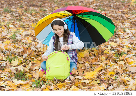 cheerful teen child relax in fall forest or park while listening music in earphones near school backpack under colorful umbrella, back to school 129188862