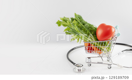 Health care banner with organic vegetables in a shopping cart with decorative heart and stethoscope on a white table with copy space. Concept of healthy food and control of good health with a diet Health care banner with organic vegetables in a shopping cart with decorative heart and stethoscope on a white table with copy space. Concept of healthy food and control of good health with a diet 129189709