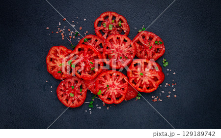 Freshly sliced ripe tomatoes arranged on a textured dark surface with herbs and salt scattered around 129189742
