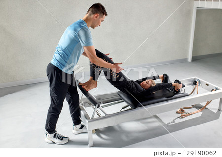 Personal trainer assisting woman on pilates reformer in modern fitness studio Personal trainer assisting woman on pilates reformer in modern fitness studio 129190062
