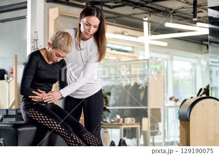 Fitness trainer assisting client during pilates session in a modern studio setting 129190075