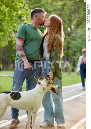 Couple sharing a kiss while walking their dog in a park setting 129190096