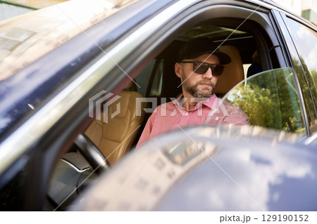 Man in a car wearing sunglasses and a hat on a sunny day 129190152