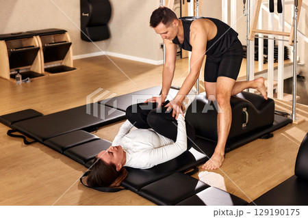 Man assisting woman in stretching exercise during a pilates session in a studio 129190175