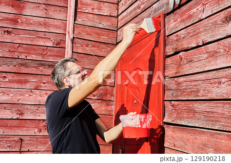House owner applies paint to metal front door using flat paint brush, man in his fifties performing home maintenance task. 129190218