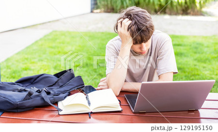 Tired of studying, a teenage boy sits with a laptop and a notebook, his head bowed in disappointment 129195150