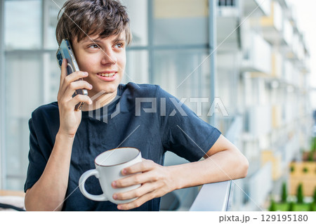 Cute teenage boy with a cup of coffee or tea happily talking on the phone on the balcony Cute teenage boy with a cup of coffee or tea happily talking on the phone on the balcony 129195180