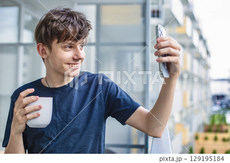 Cute teenage boy with a cup of coffee or tea happily talking on the phone on the balcony Cute teenage boy with a cup of coffee or tea happily talking on the phone on the balcony 129195184