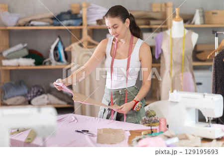 Attentive young female dressmaker holding textile detail fixed on paper 129195693