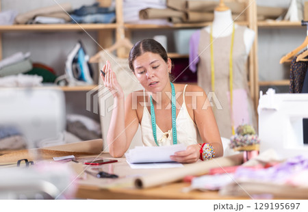 Young tailoress engaged in paperwork in sewing workshop 129195697