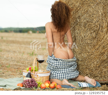 Naked woman in open shirt kneeling by haystack during secluded picnic 129195752