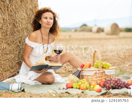 Woman sitting on ground in field, settled down in nature with book and glass of wine 129195763