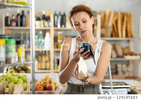 Female shopper scans QR code on cheese label using smartphone. Checking products in supermarket Female shopper scans QR code on cheese label using smartphone. Checking products in supermarket 129195883