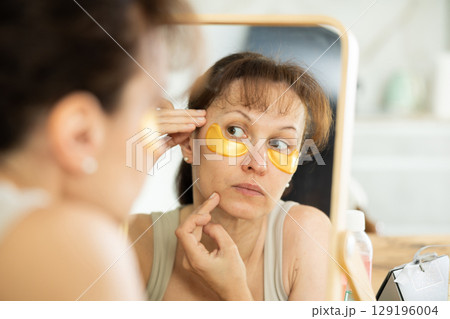 Middle-aged woman applying eye patch mask on her face sitting in front of the mirror Middle-aged woman applying eye patch mask on her face sitting in front of the mirror 129196004