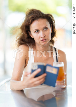 Woman with loose hair reading information on smartphone screen and drinking beer in open air restaurant Woman with loose hair reading information on smartphone screen and drinking beer in open air restaurant 129196053
