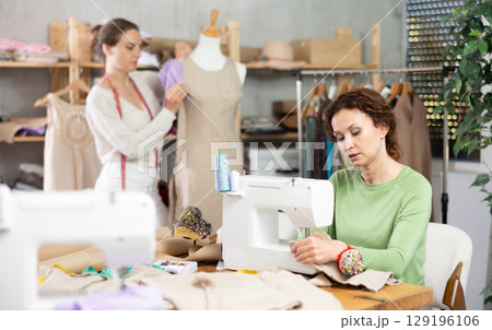 Working in sewing workshop - female tailor sewing on sewing machine, another tailor trying on dress on mannequin 129196106