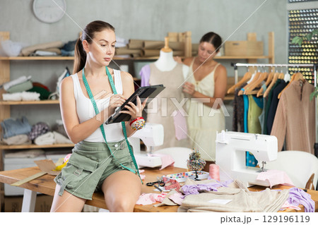 Female tailor create sketch of dress on tablet. Assistant work with mannequin in background Female tailor create sketch of dress on tablet. Assistant work with mannequin in background 129196119