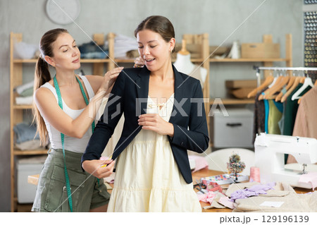 Young tailoress trying ready jacket on female client Young tailoress trying ready jacket on female client 129196139