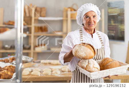 Experienced aged woman in apron working in bakery kitchen, posing with baked bread 129196152