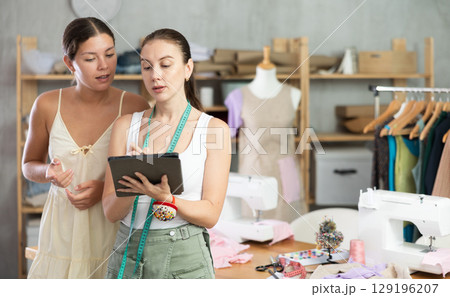 Woman tailor and girl client discuss manufacture of clothes, look at tablet screen 129196207
