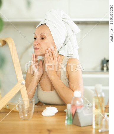 Woman with hair wrapped in towel inspecting complexion in mirror Woman with hair wrapped in towel inspecting complexion in mirror 129196259