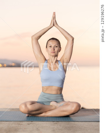 Portrait of caucasian woman sitting on sports mat and making yoga meditation in lotus pose and hands up on seashore at sunrise Portrait of caucasian woman sitting on sports mat and making yoga meditation in lotus pose and hands up on seashore at sunrise 129196276