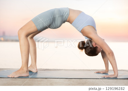 Portrait of caucasian experienced woman yoga teacher performs ardha chakrasana on seashore at sunset Portrait of caucasian experienced woman yoga teacher performs ardha chakrasana on seashore at sunset 129196282