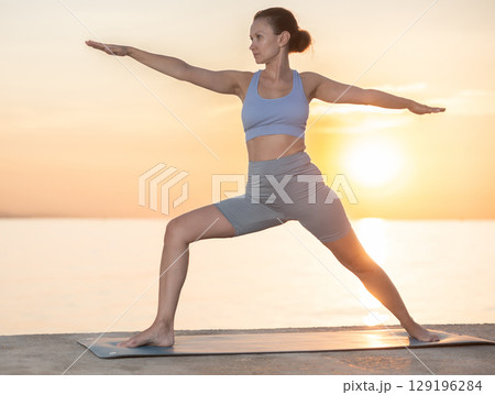 Happy woman coach standing yoga pose and making full body stretching workout on seashore at sunrise Happy woman coach standing yoga pose and making full body stretching workout on seashore at sunrise 129196284